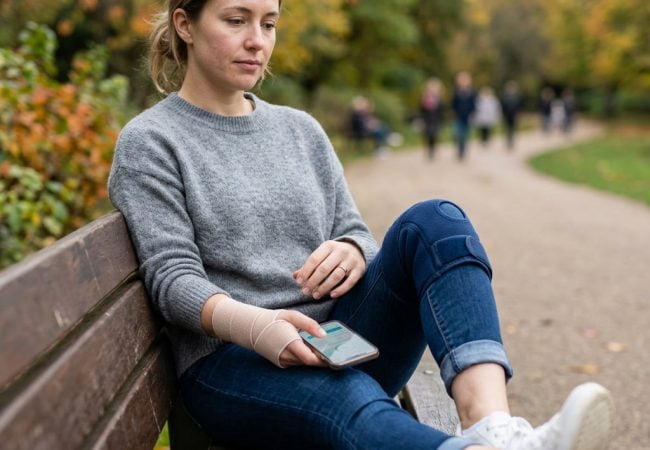A woman with wrist and knee braces sits on a park bench, holding a smartphone, with autumn trees and people walking in the background.
